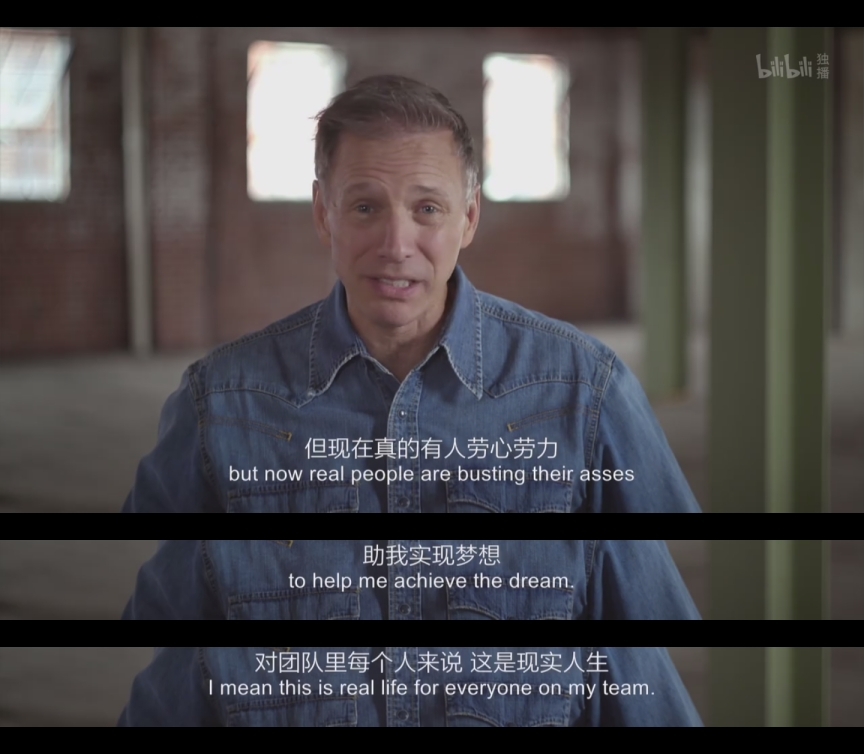 一位男性在空旷的室内讲述他的创业经历,身着牛仔衬衫,背景是模糊的立柱和窗户,表情严肃而投入。