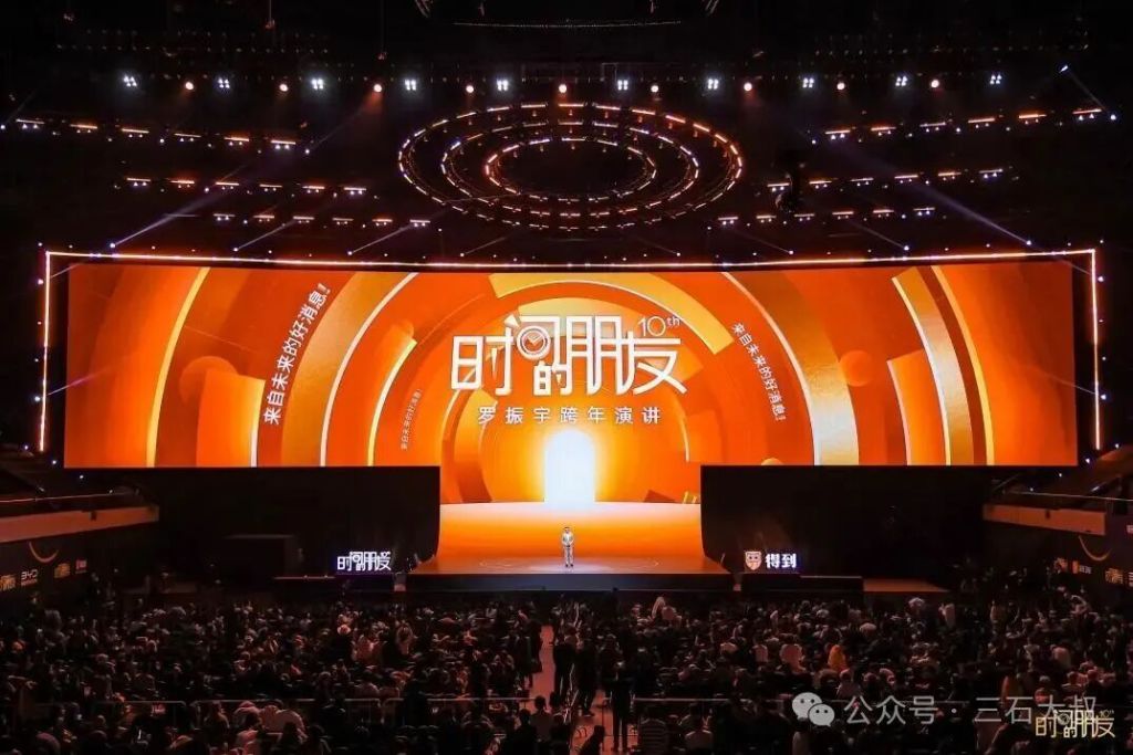 一个大型会场，舞台上有橙色主题背景和大型屏幕，显示着"时尚开放"和"罗振宇年度演讲"的字样，观众席满是观众，形成热烈的场面。